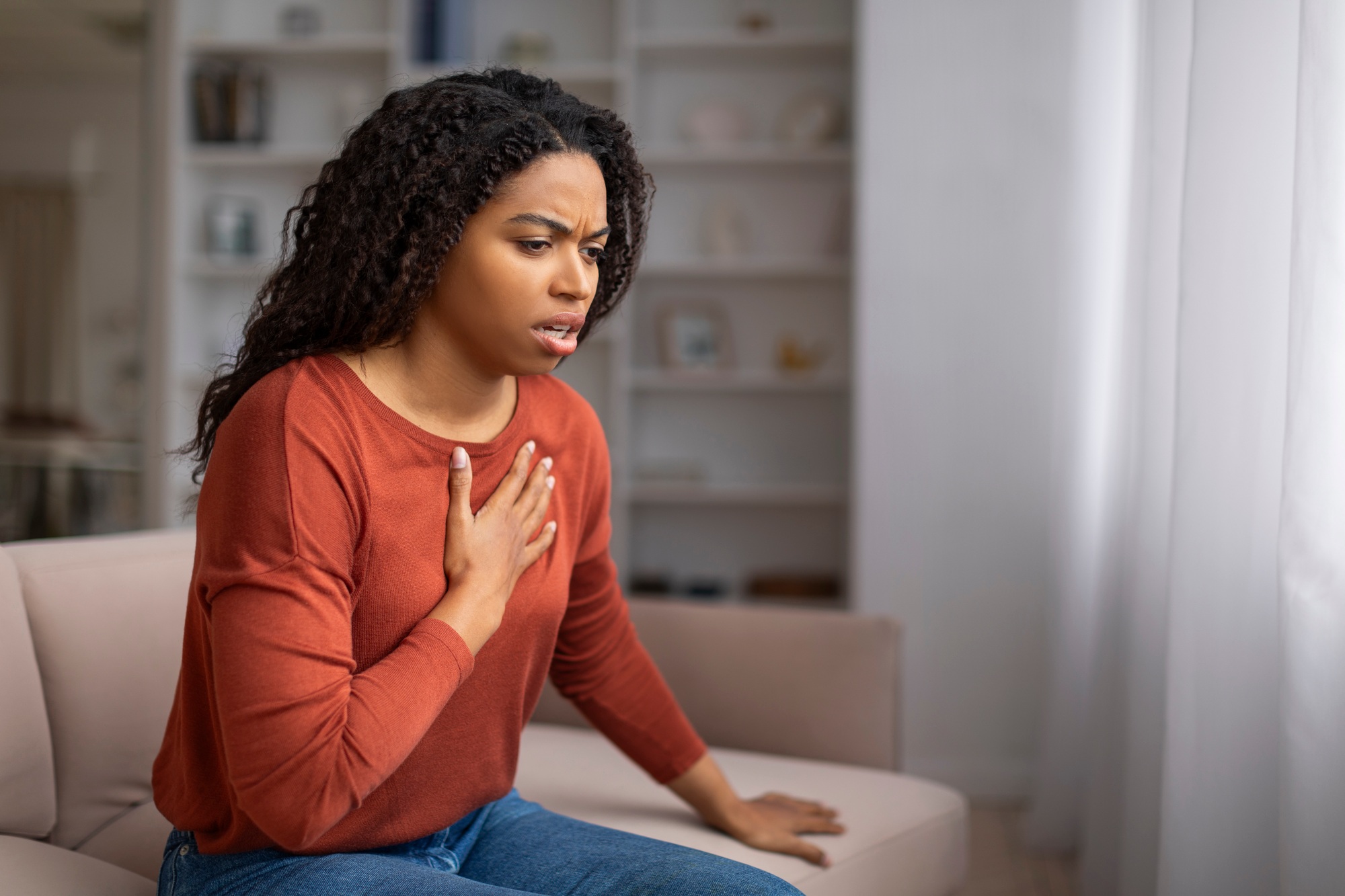 Concerned young black woman suffering pain in chest while sitting on couch